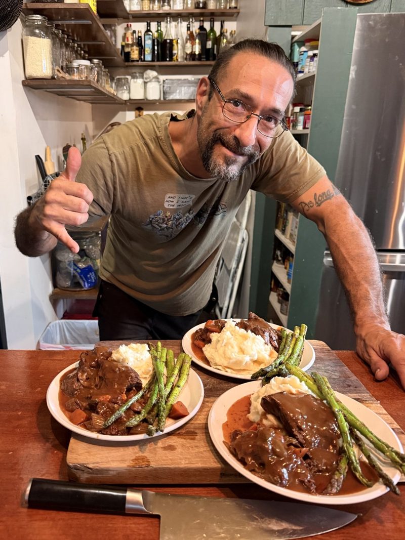 Chef Anthony Rinaldi in his kitchen, smiling and giving a shaka behind three plates of beef with gravy, mashed potatoes, and asparagus, with a chef's knife on the counter.
